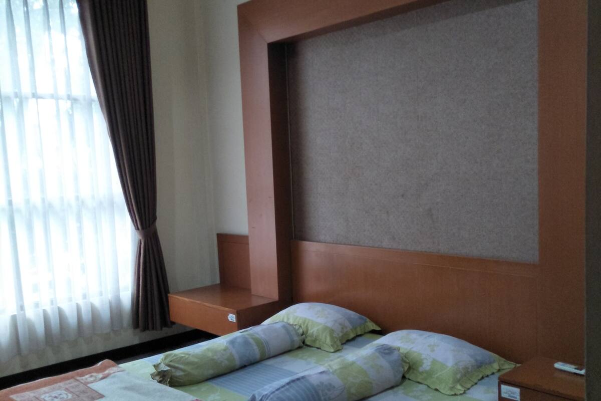 Ein Schlafzimmer mit einem Bett, einem Kopfteil, einem Nachttisch und einem Fenster mit Vorhängen.