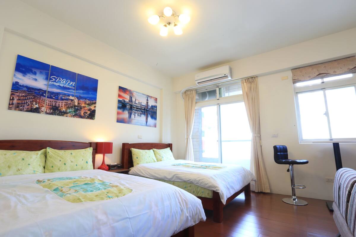 A hotel room with two beds, a window with curtains, an air conditioning unit, and a cityscape poster on the wall.