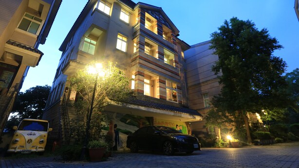 A multi-story building with a balcony on the second floor, a car parked in front, and a tree to the right. - Downshifted House (Miaoli)