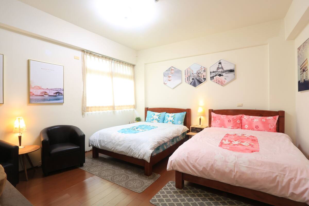 A hotel room with two beds, a chair, a desk, and a lamp.