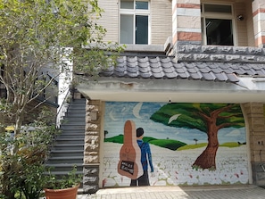 A building with a mural of a tree and a person, a staircase, and a potted plant. - Downshifted House (Miaoli)