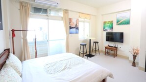 A hotel room with a bed, a TV, a window with curtains, a wall-mounted air conditioner, and a small table with a vase of flowers. - Downshifted House (Miaoli)