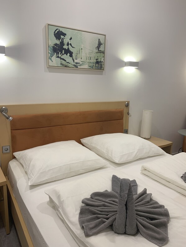 Una cama bien hecha con ropa de cama blanca y una toalla gris, un cabecero de madera, una mesita de noche con una lámpara y un cuadro colgado en la pared.