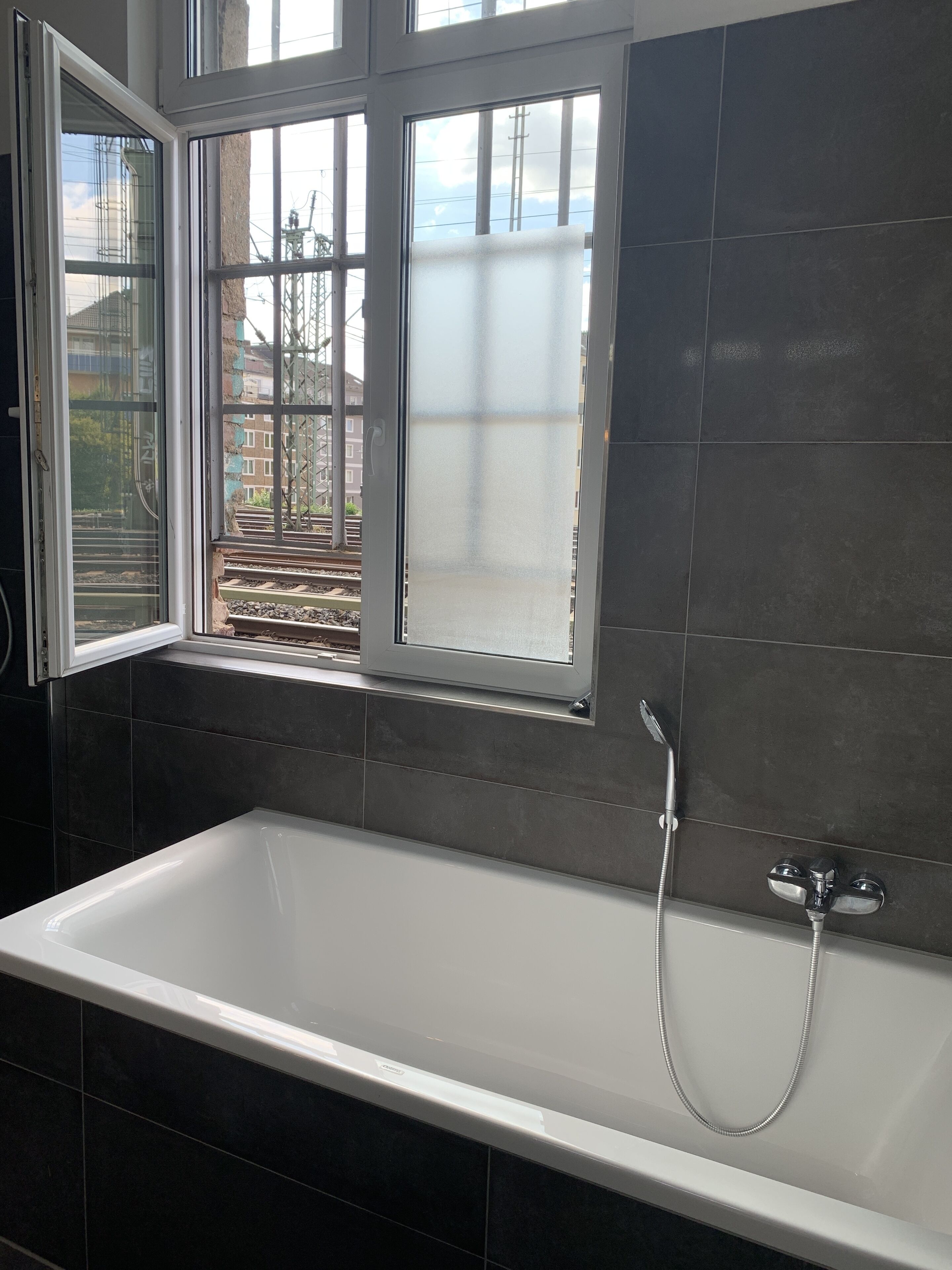 A modern bathroom with a large bathtub, a window with a view of buildings and a train track, and dark tiled walls.
