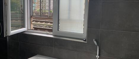 A modern bathroom with a large bathtub, a window with a view of buildings and a train track, and dark tiled walls.