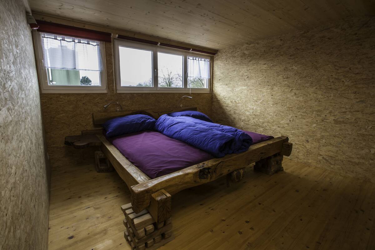 A wooden bed in a room with a window and a view of the outside.
