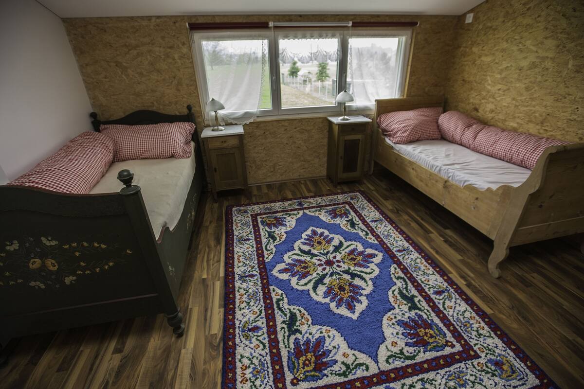 A room with two beds, a window with curtains, a wooden floor, and a patterned rug.