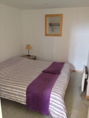 Une chambre avec un lit simple, une couvre-lit à rayures, une couverture violette, une lampe de chevet et un miroir avec un cadre en bois accroché au mur.