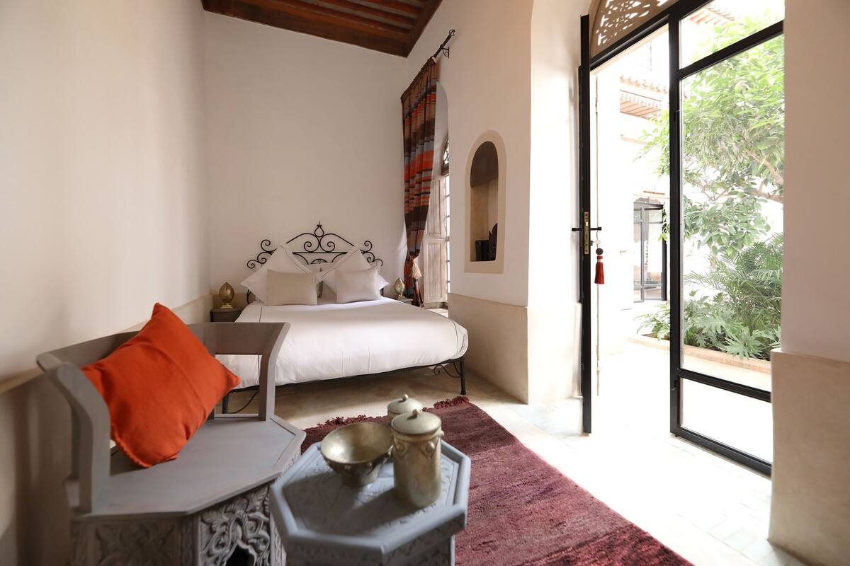 A bedroom with a bed, a nightstand, a bedside table with a lamp, and a view of the outdoors through a large window.