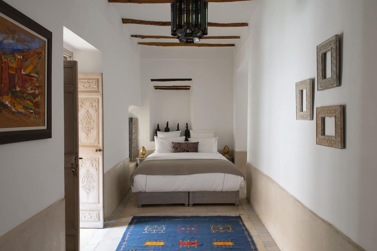A narrow hallway leading to a bed with a headboard, a blue patterned rug, and framed pictures on the walls.