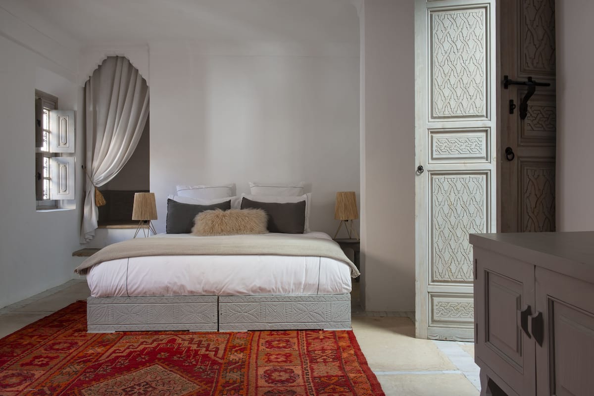 A bedroom with a bed, curtains, a nightstand, a lamp, a wardrobe, and a patterned rug.