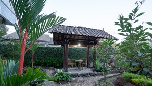 A covered outdoor seating area with a tiled roof, surrounded by lush greenery and a stone pathway.