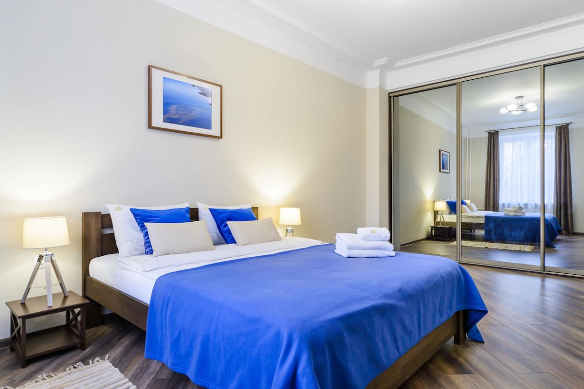 A hotel room with a large bed, two bedside tables with lamps, a framed picture on the wall, and a large mirror reflecting the room.