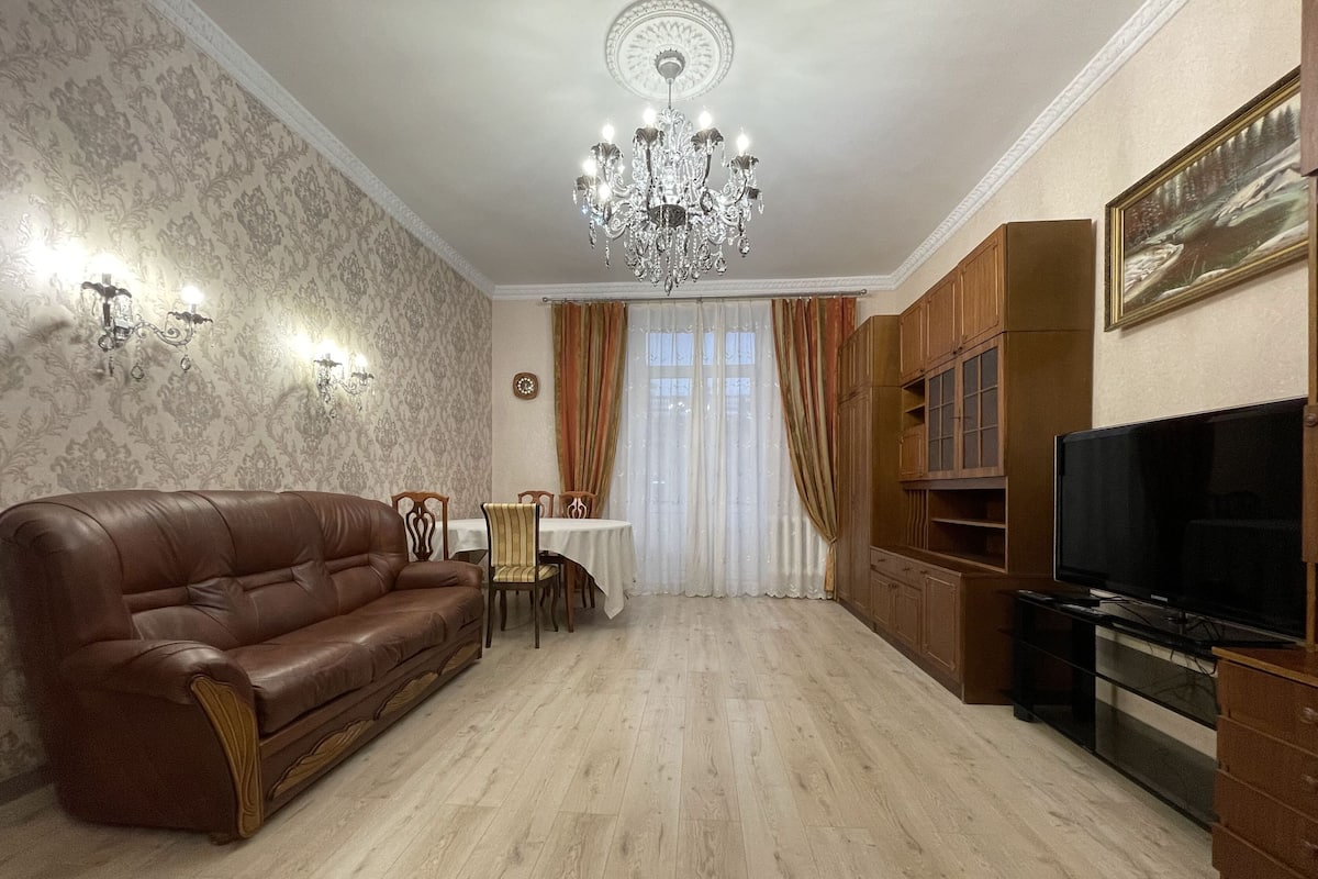Ein Wohnzimmer mit braunem Ledersofa, einem Esstisch mit Stühlen, einer Deckenleuchte, Holzschränken und einem Fernseher.