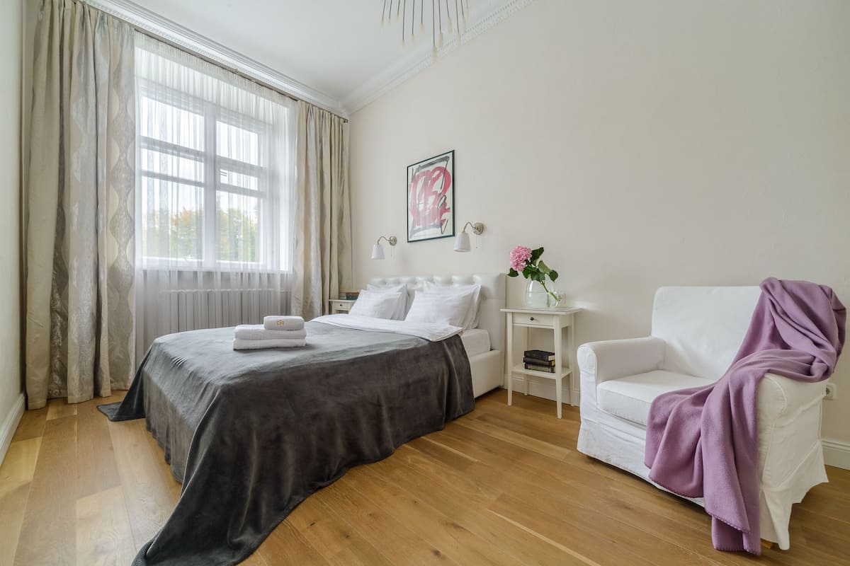 Ein Schlafzimmer mit einem Bett, einem weißen Stuhl, einem Nachttisch und einem Fenster mit Vorhängen.