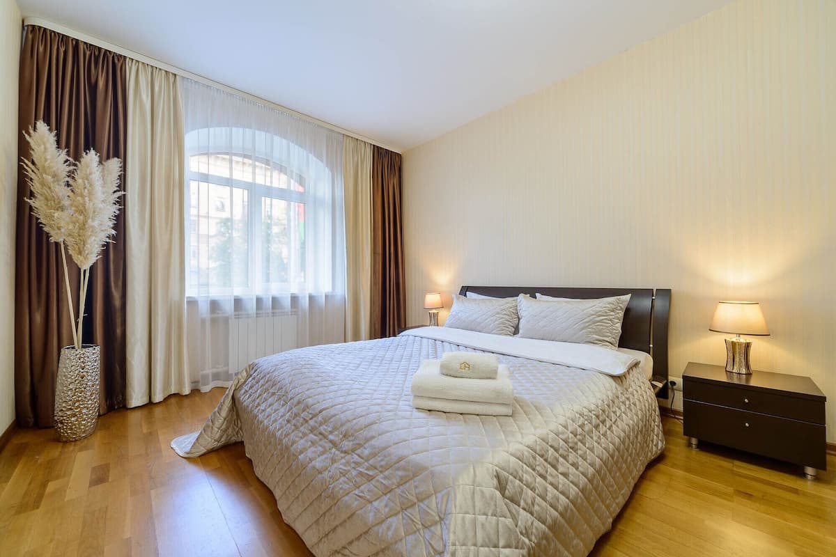 Ein ordentlich bezogenes Bett mit gesteppter Bettdecke, ein Nachttischchen mit Lampe und ein Fenster mit durchscheinenden Vorhängen und einer dekorativen Vase mit Pampasgras.
