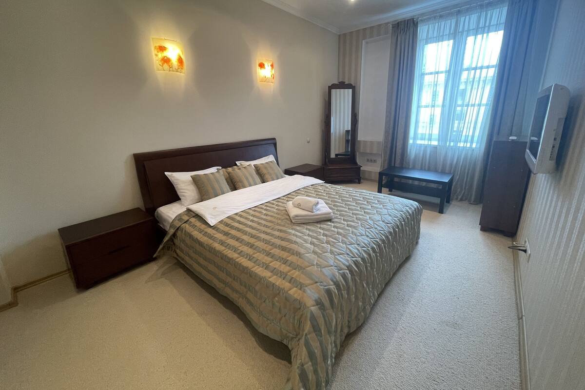 A neatly arranged hotel room with a large bed, bedside tables, a mirror, a TV, and a window with curtains.