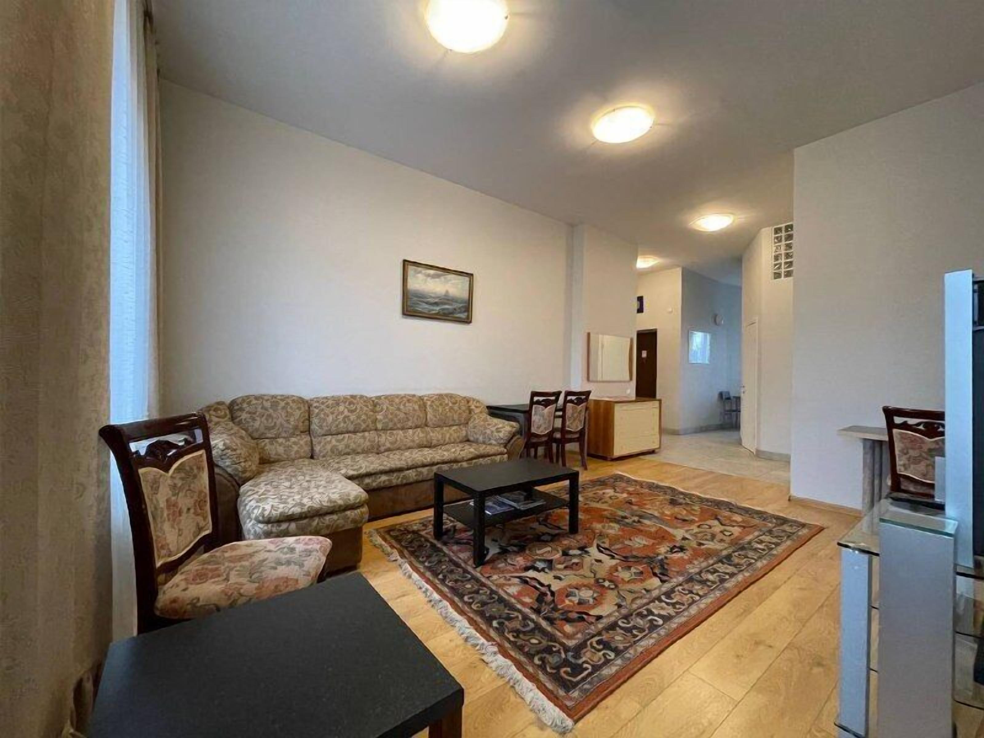 A living room with a sofa, chairs, a coffee table, and a rug. There is a painting on the wall and a door leading to another room.