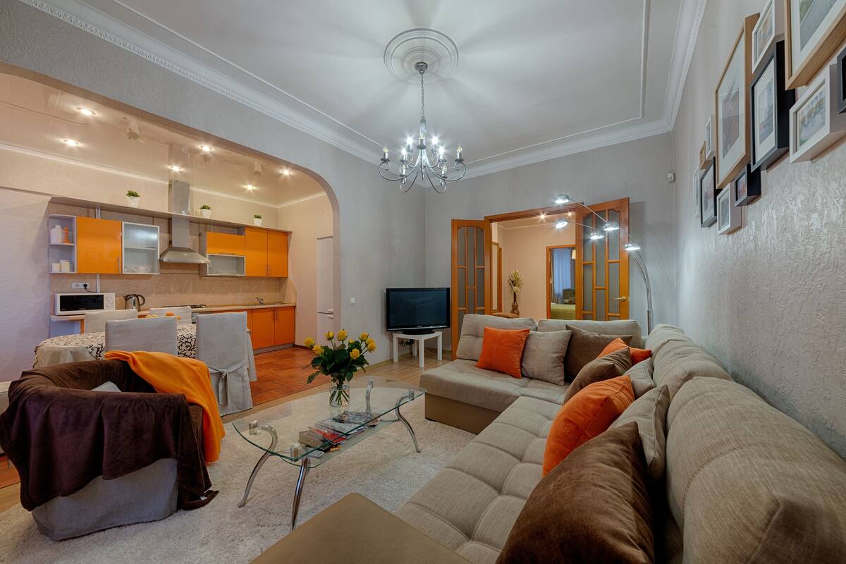 A modern living room with a sofa, armchairs, a glass coffee table, and a TV. There is a kitchen area with cabinets and a microwave.