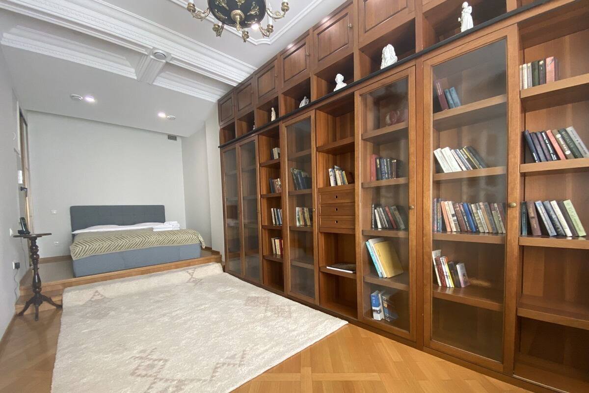 A bedroom with a large bed, a wooden wardrobe with glass doors, and a bookshelf filled with books.