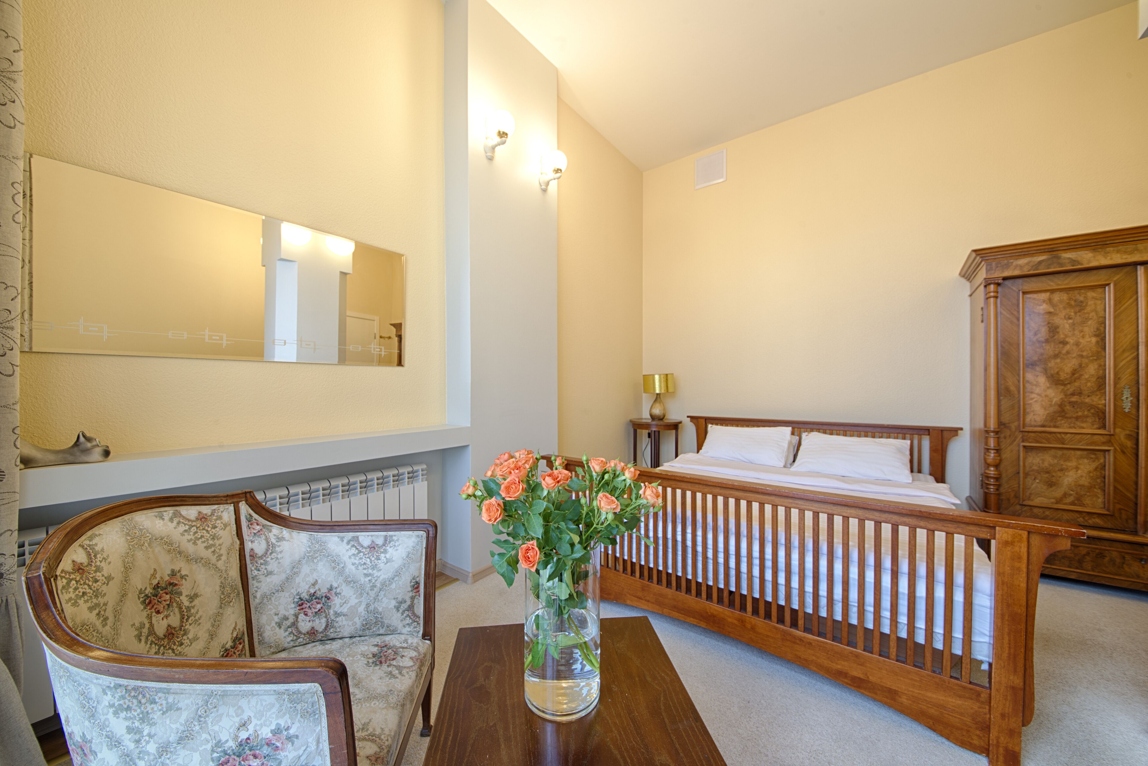 Une chambre avec un lit en bois, une chaise, une table ornée d’un vase de fleurs et un grand miroir accroché au mur.
