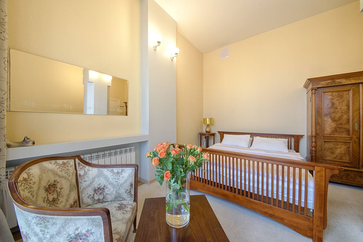 A bedroom with a wooden bed, a chair, a table with a vase of flowers, and a large mirror on the wall.