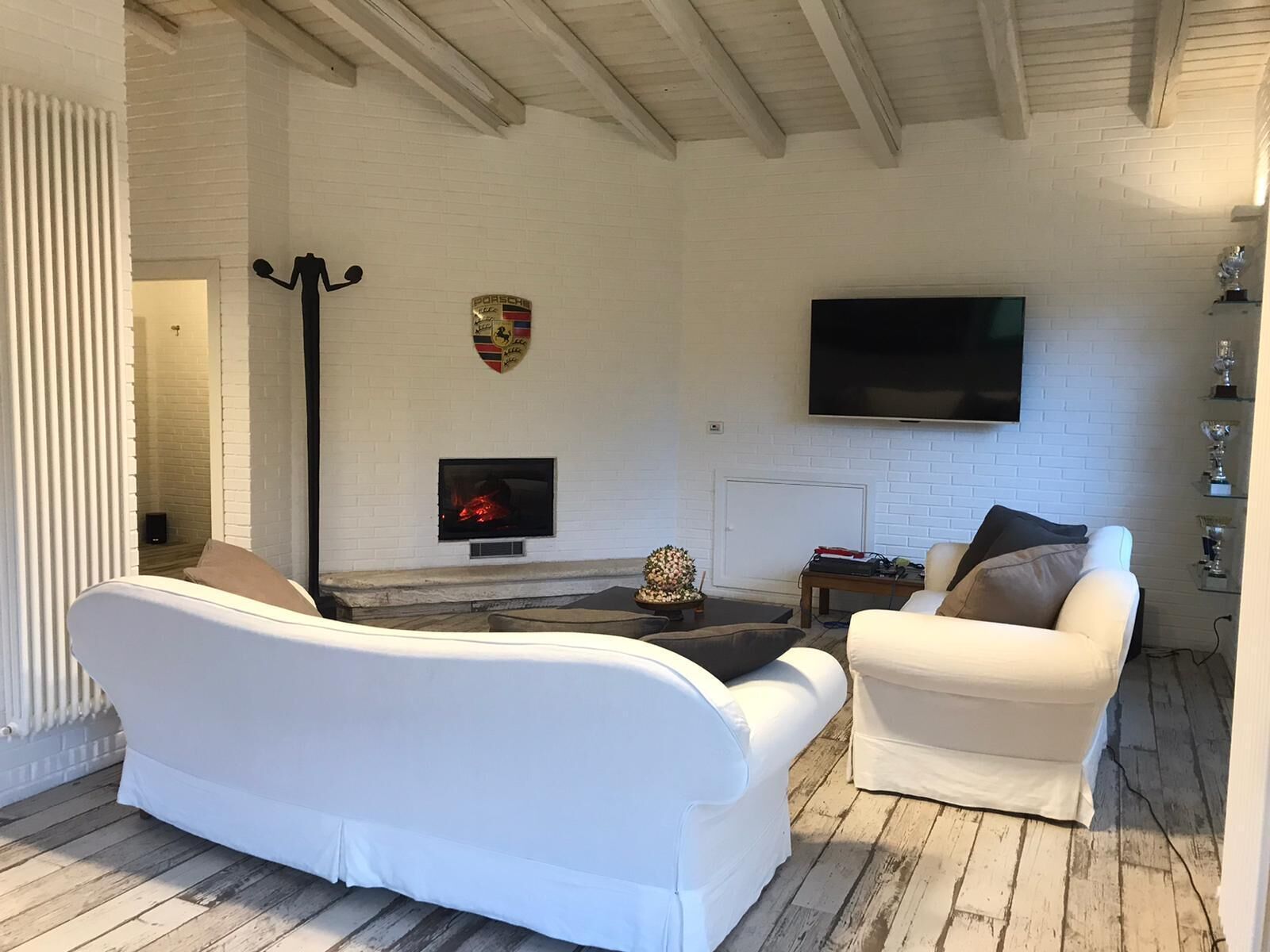 A living room with a white sofa, a white armchair, a fireplace, a flat-screen TV, and a decorative shield on the wall.