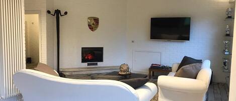 A living room with a white sofa, a white armchair, a fireplace, a flat-screen TV, and a decorative shield on the wall.