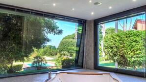 Een moderne badkamer met een groot bad, grote ramen en uitzicht op een zwembad en groen.