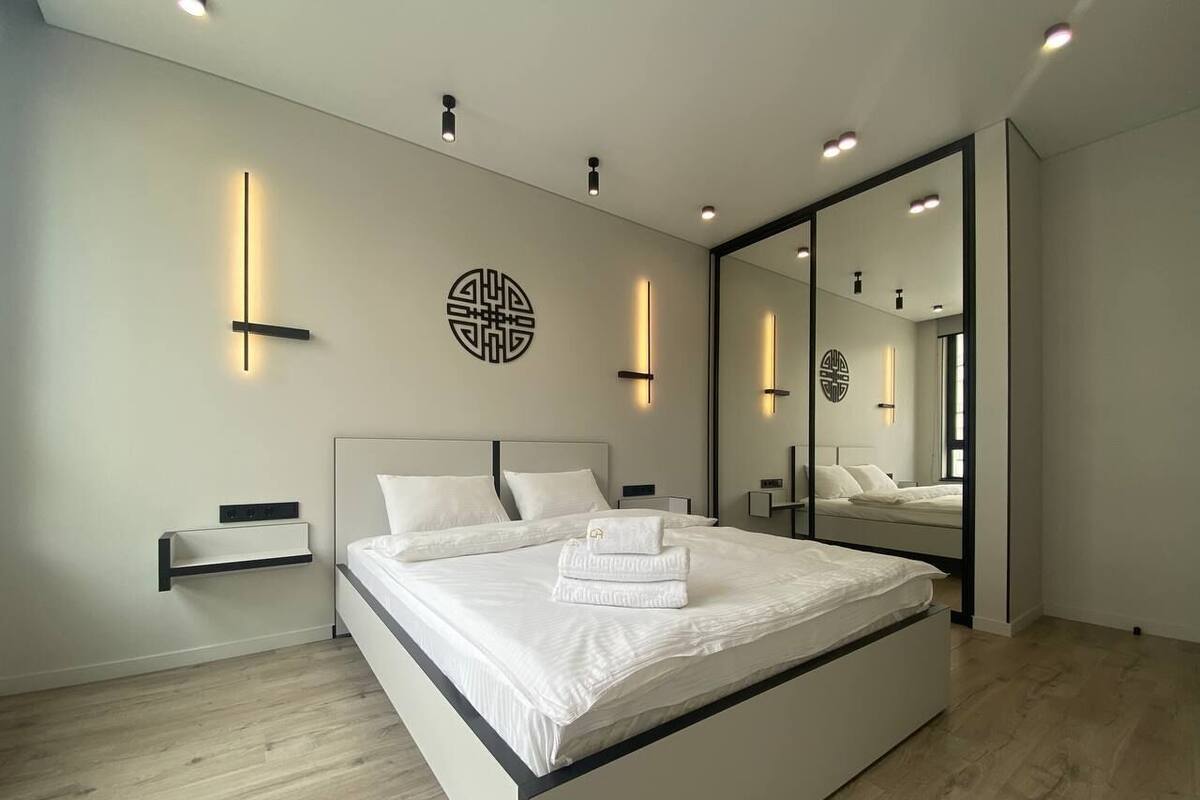 A modern bedroom with a large bed, bedside tables, wall-mounted lighting, and a mirror.