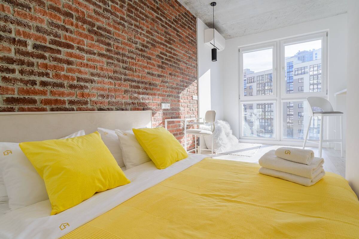 A modern hotel room with a brick wall, a large bed with yellow bedding, and a view of buildings through large windows.