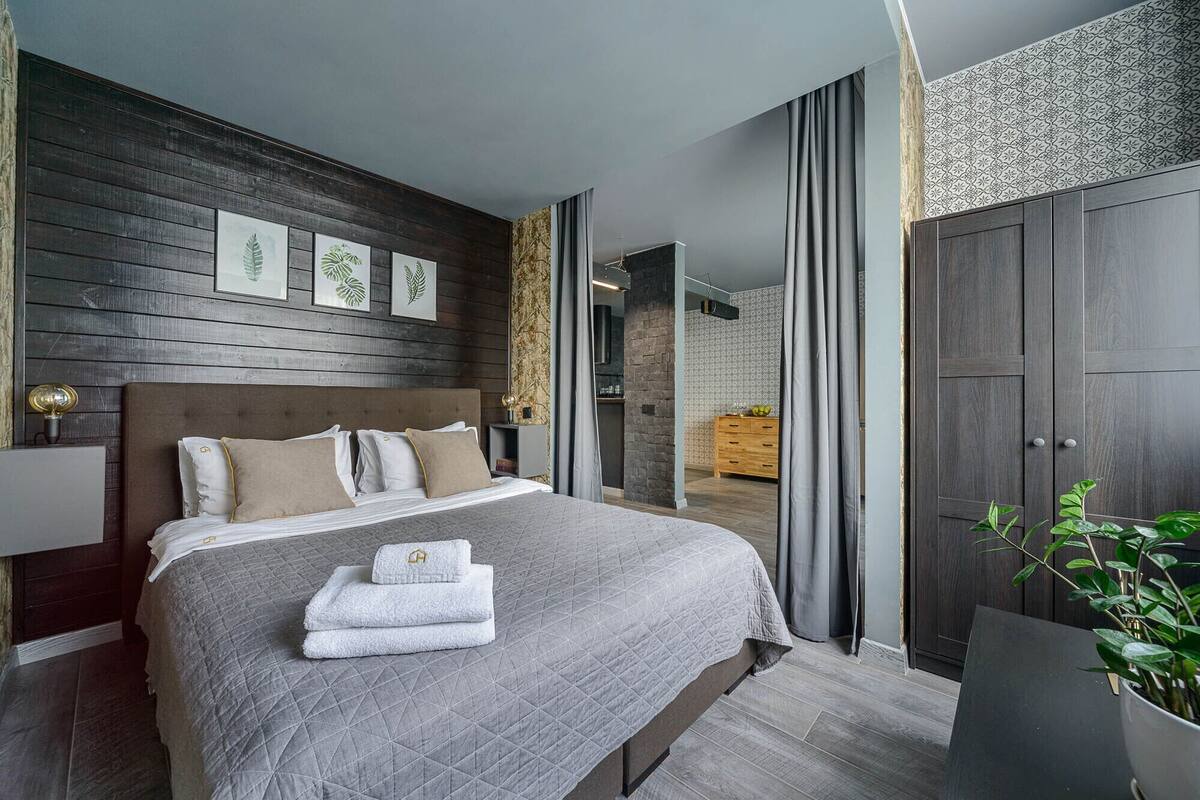 A modern bedroom with a large bed, a wooden headboard, and a dark wall with leaf-patterned artwork.