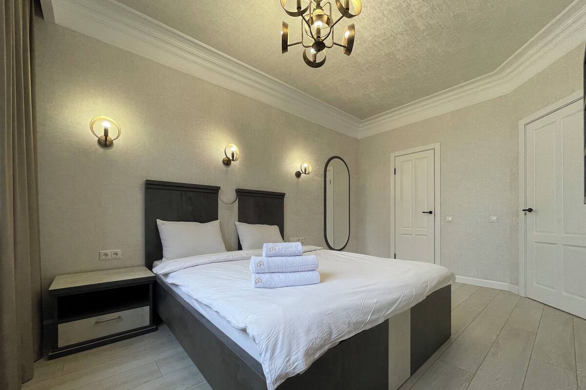 A modern bedroom with a large bed, bedside tables, a mirror, and a chandelier.