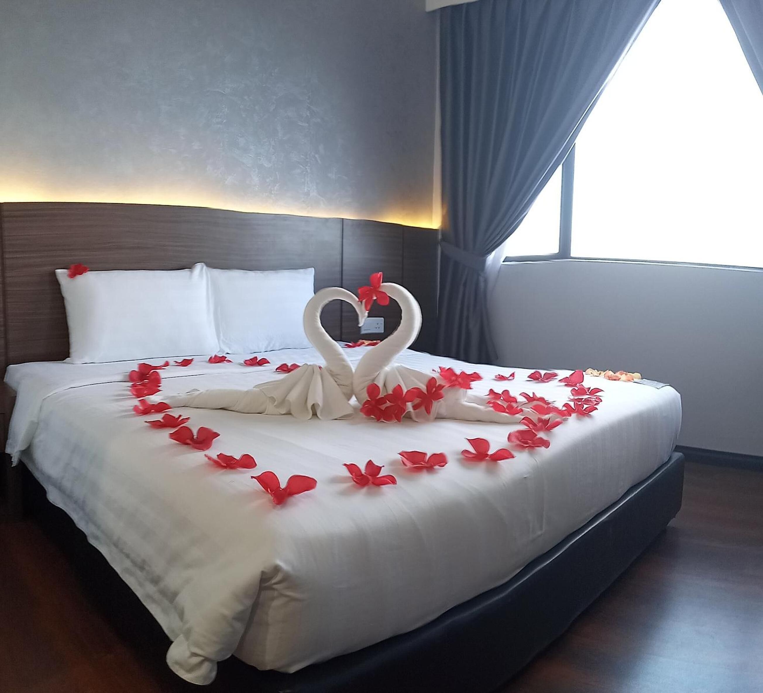 A neatly made bed with a heart-shaped decoration made of flowers and a swan figure, surrounded by red flowers.