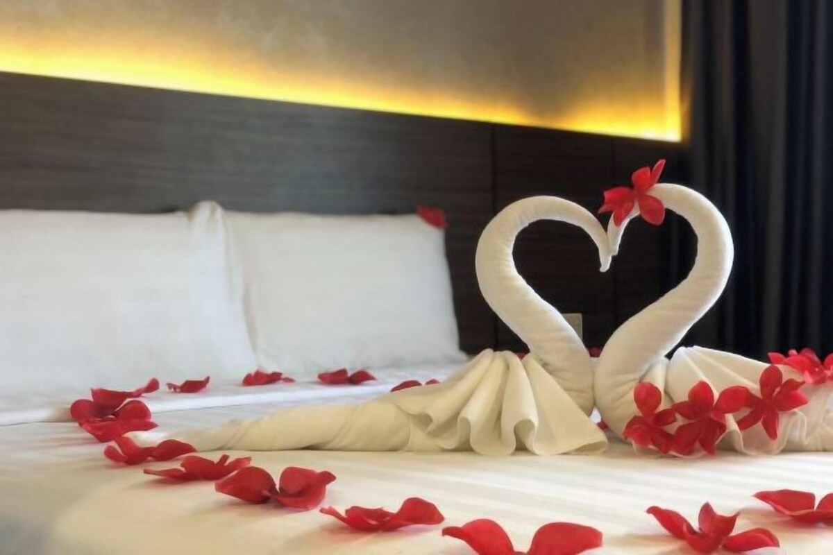 A bed with a white sheet, two swans made of towels, and red flowers scattered on the bed.