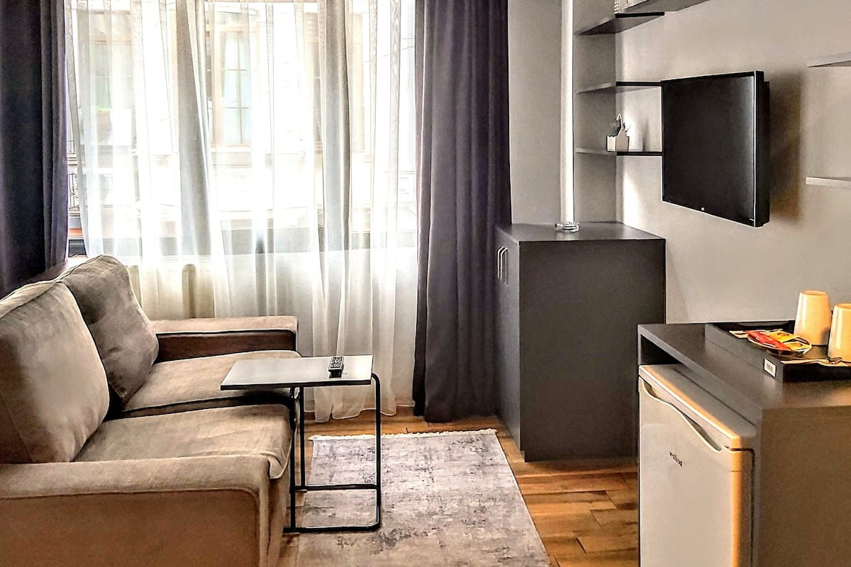 A modern living room with a sofa, a small table, a TV mounted on the wall, and a kitchen area with a refrigerator and microwave.