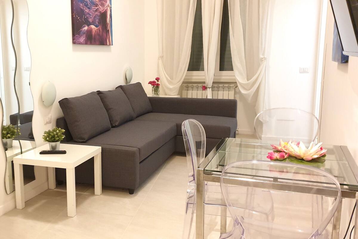 A modern living room with a grey sofa, a white side table, and a glass dining table.