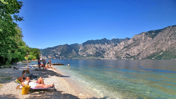 A beach with clear water, pebbles, and mountains in the background. - Hotel Baitone (Malcesine)