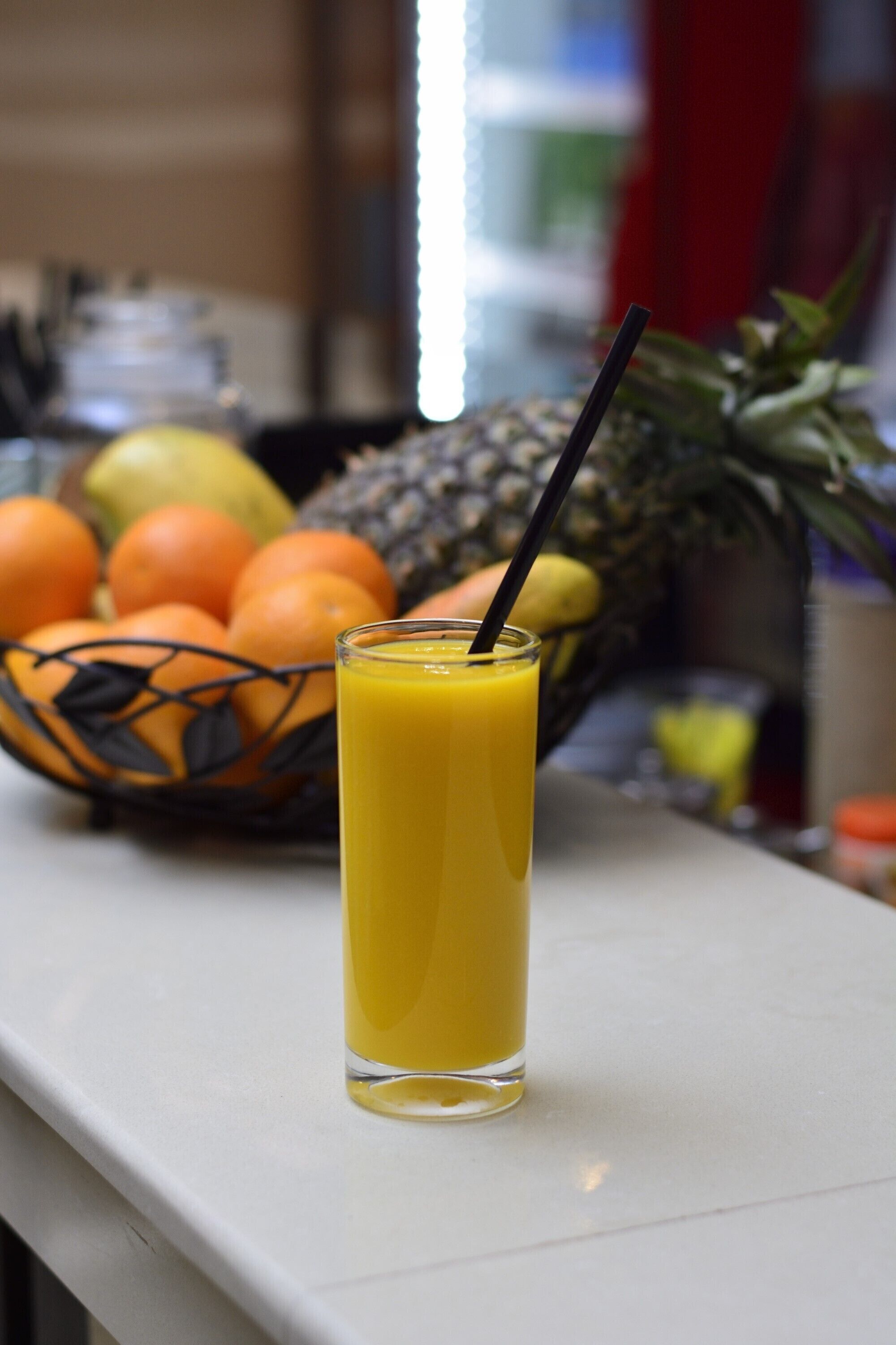 A glass of orange juice with a straw on a white countertop, with a basket of oranges and a pineapple in the background.