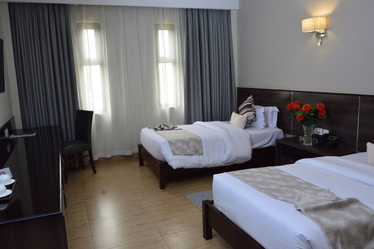 A hotel room with two beds, a desk, a chair, a nightstand with a vase of flowers, and a wall-mounted lamp.