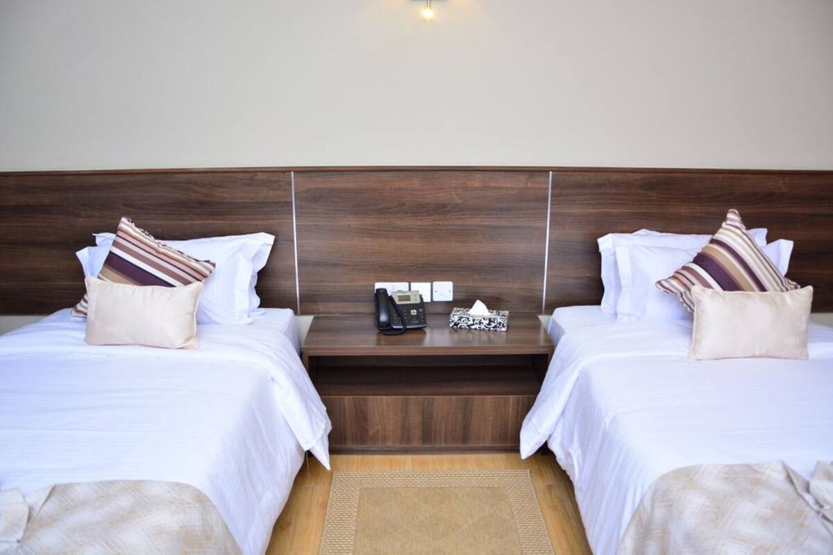 A hotel room with two beds, a wooden headboard, a bedside table with a phone and a tissue box, and a small rug.