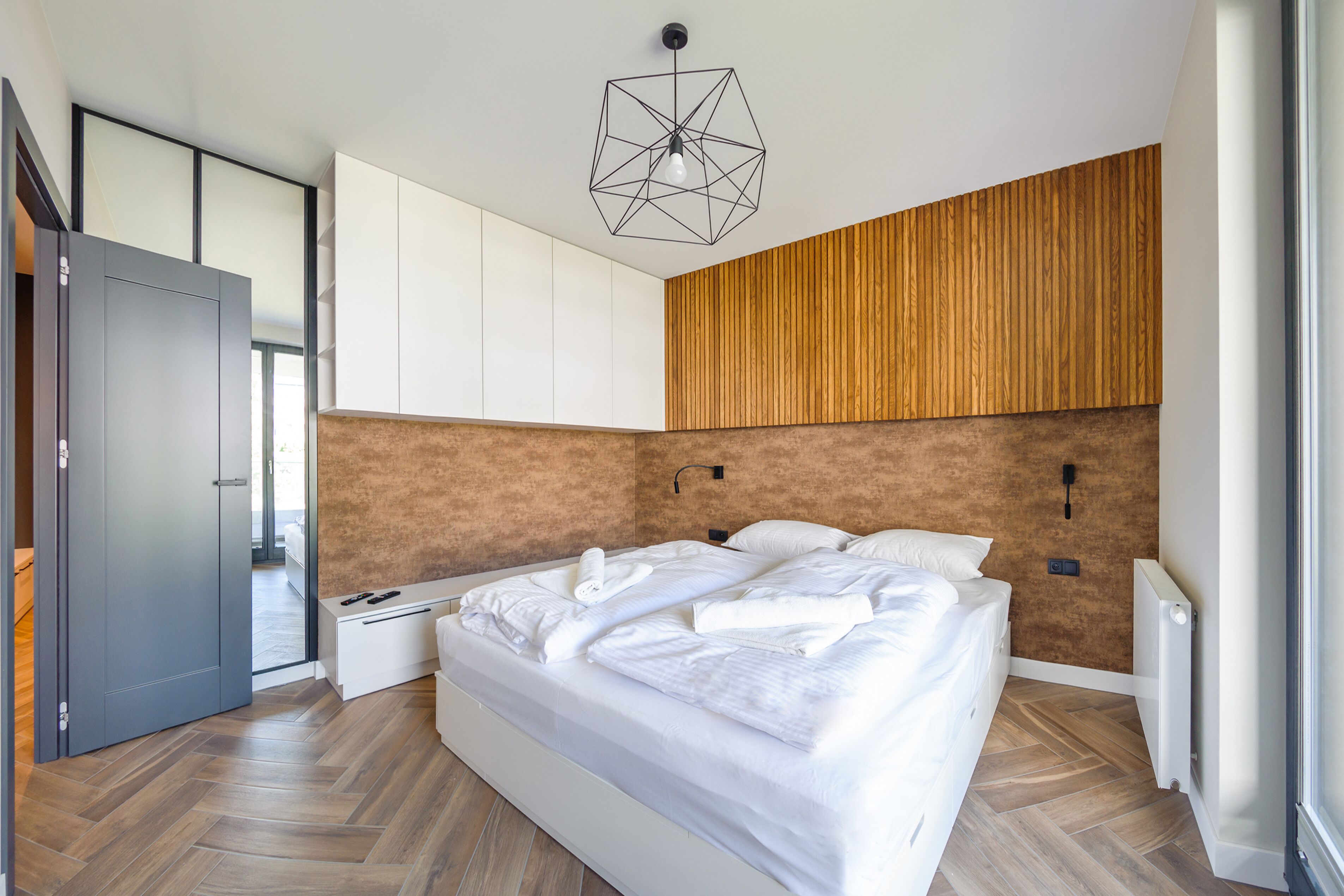 A modern bedroom with a large bed, wooden panel wall, and a geometric light fixture.
