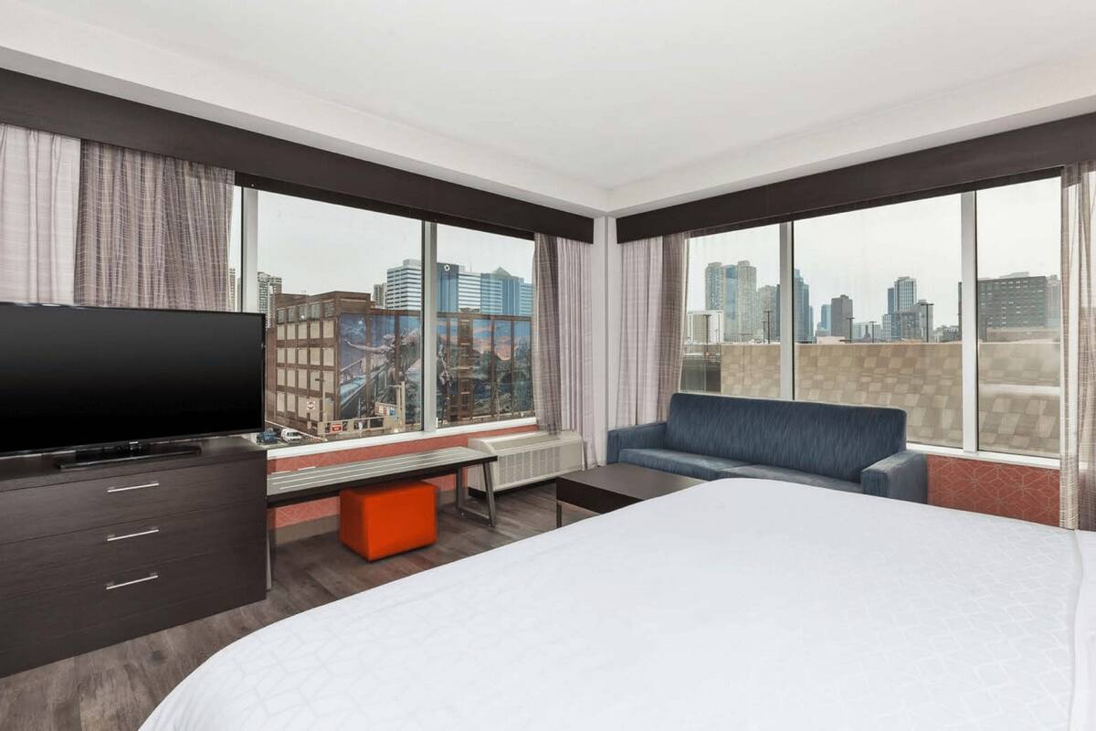 Ein Hotelzimmer mit einem großen Bett, einem Fernseher, einer Couch und Blick auf die Stadt durch große Fenster.