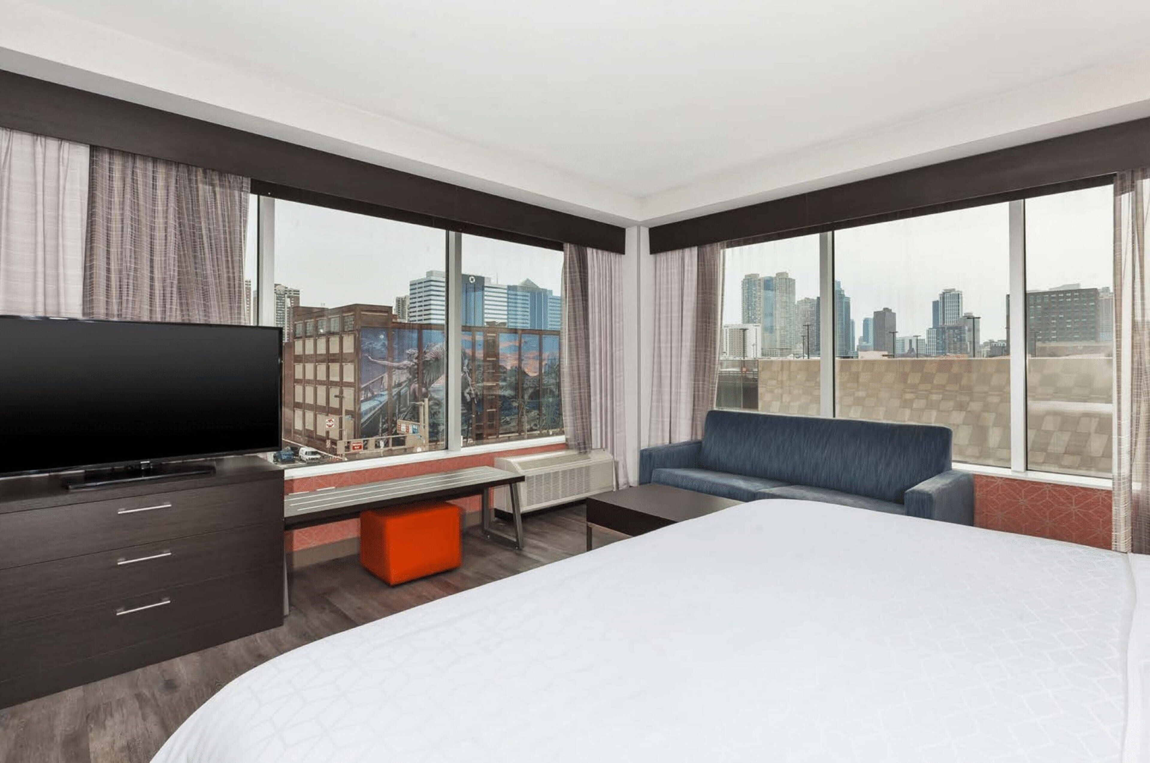 A hotel room with a bed, a TV, a sofa, and large windows with a city view.