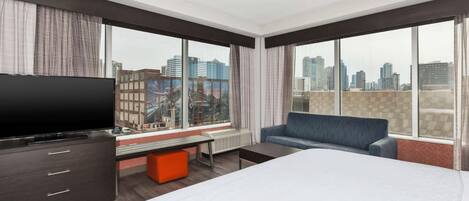 A hotel room with a bed, a TV, a sofa, and large windows with a city view.