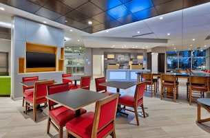 Un interior de restaurante moderno con asientos tapizados en rojo y mesas de madera, un televisor de pantalla plana grande montado en la pared y vistas de la ciudad a través de amplias ventanas.