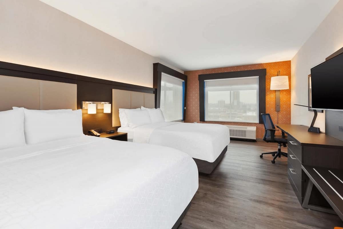 Ein Hotelzimmer mit zwei Betten, einem Schreibtisch, einem Sessel, einem Fernseher und einem großen Fenster mit Blick auf die Stadt.