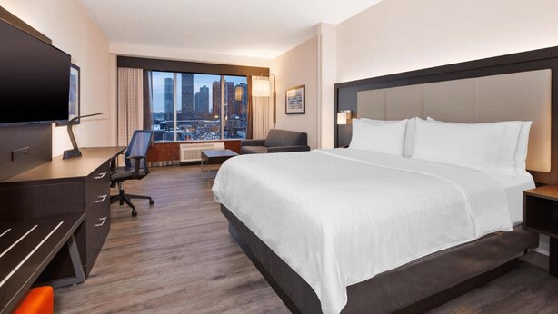 A hotel room with a large bed, a desk, a chair, a TV, and a city view.