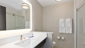 A modern bathroom with a large mirror, a white sink, a glass shower enclosure, and a toilet with towels hanging on the wall.