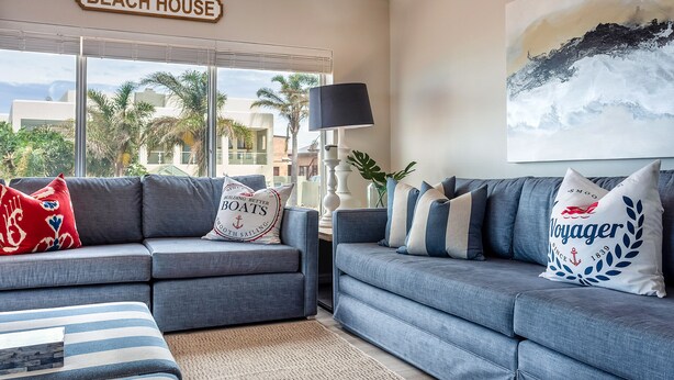 A living room with two grey sofas, a striped ottoman, and a 'Beach House' sign above the window. - Villa Nikos J-Bay (Jeffreys Bay)